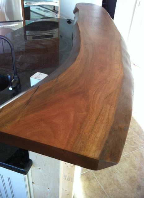 Thick live-edge African mahogany kitchen counter curving along a black granite island, rich reddish-brown grain with a natural undulating edge.