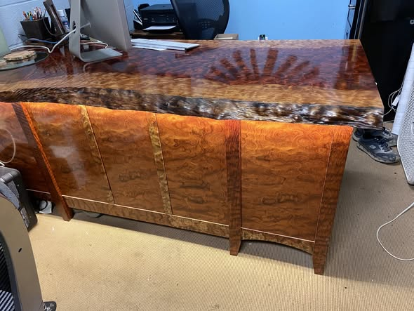 Live-edge bubinga office desk with mirror-gloss finish, dramatic figured grain and a sunburst medallion at one end, sitting above a matching bubinga case piece with four burl-front panels and arched apron.