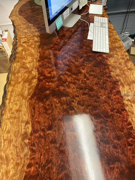 Top-down view of the live-edge bubinga desk with iMac and keyboard, showing wavy live edge and chatoyant burl figure under glassy finish.