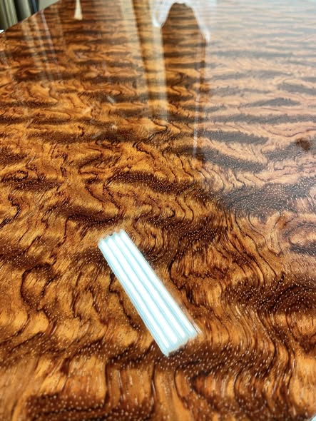 Close-up of figured bubinga grain under a glassy mirror finish, swirling chatoyant pattern with light reflections.