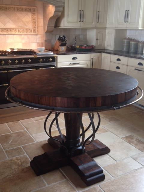 Round end-grain butcher block table on a heavy timber pedestal base, wrapped with a hand-distressed metal band that incorporates an integrated towel bar, set on travertine tile in a French country kitchen.