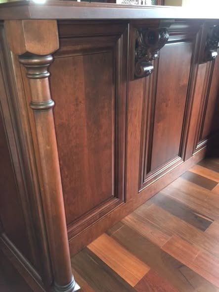 Close-up of the cherry home bar showing the turned column, raised panel doors, carved corbel above, and applied moldings on the lower rail.