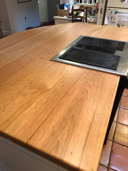 American cherry kitchen island countertop with wide plank boards, warm pinkish-amber tone and low-sheen clear finish, with a stainless downdraft grill cooktop set into the surface, terracotta tile floor below.