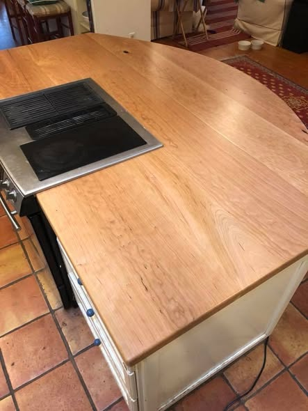 Opposite-end view of the cherry island top showing the rounded end, wide boards, cooktop on the left, and painted island base set on Saltillo tile.