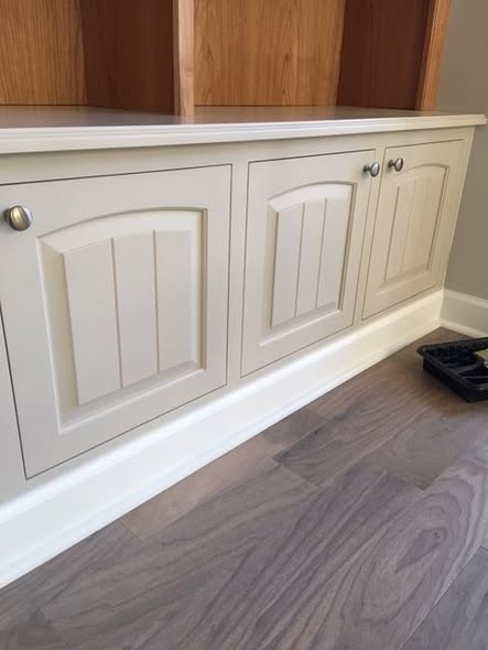 Close-up of the painted base cabinet beneath the cherry bookcase, featuring arched raised panel doors with a beaded face frame on a walnut floor.