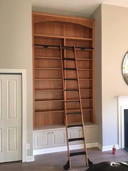 Full-room view of the 12-foot cherry bookcase with rolling ladder beside a fireplace, showing the painted base cabinets and arched cherry top.