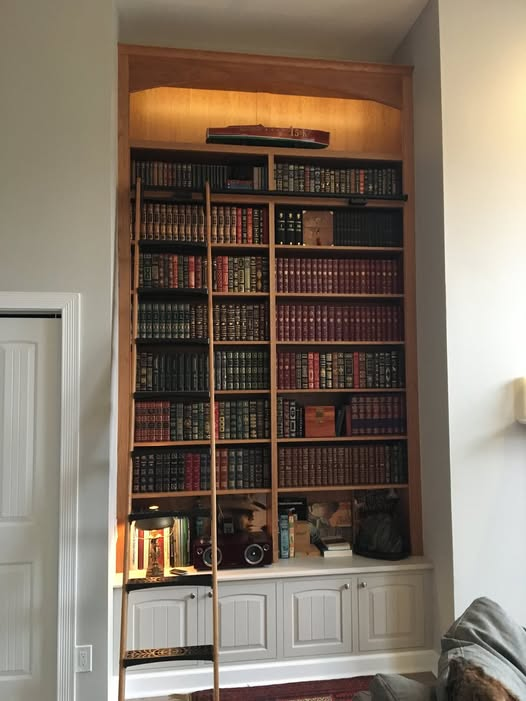 Floor-to-ceiling cherry library bookcase loaded with leather-bound volumes, integrated rolling library ladder, arched valance with interior lighting, and painted raised-panel base cabinet below