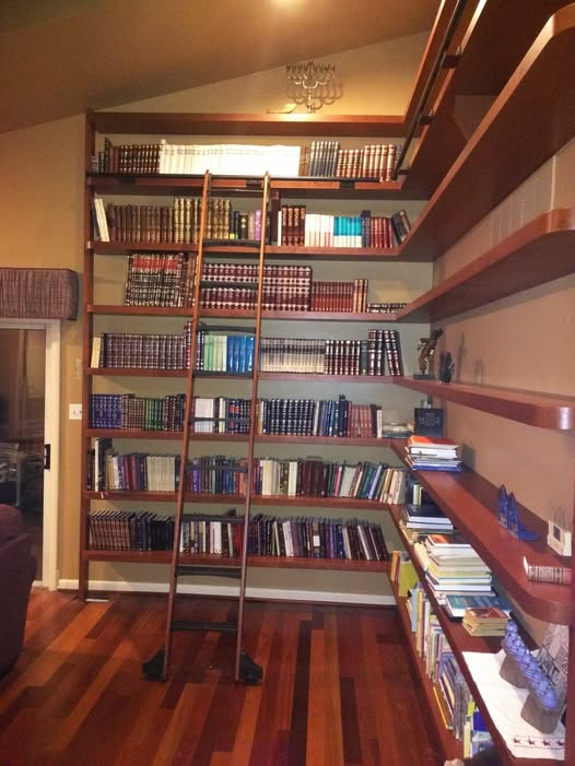 Floor-to-ceiling cherry floating shelves filled with books wrapping around a corner, with a rolling library ladder on a brass rail and Brazilian cherry hardwood floors.