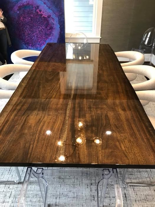 A 9-foot long live edge dining table with a rich figured wood top and high-gloss multilayered clear coat reflecting overhead lights, surrounded by sculptural cream chairs and clear acrylic chairs.
