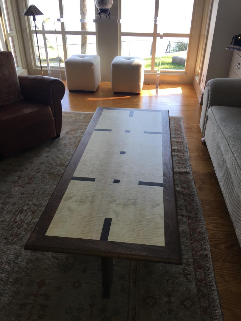 Rectangular coffee table with a figured curly maple top bordered in walnut, accented with dark walnut inlay strips in a geometric pattern, sitting on a patterned rug in a sunlit living room.