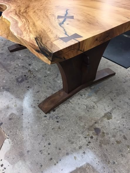 Corner detail of the live-edge Matumi slab showing the walnut bowties, thick top, natural crack detail, and sculpted walnut trestle base below.