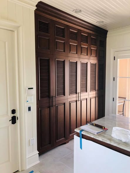 Tall white oak mudroom cabinets stained a rich dark brown, featuring a row of 9-foot doors with louvered upper panels and raised lower panels, designed to look like three doors but open as one.
