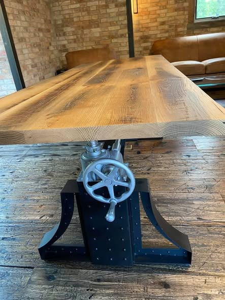 Reclaimed white oak desk top with rich amber tones and visible saw marks, resting on an antique cast-iron crank-adjust base with a spoked hand wheel, set on a wide-plank wood floor inside a brick-walled room.