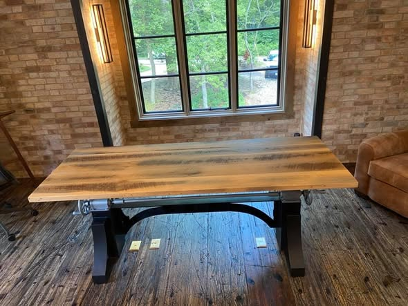 Front view of the reclaimed white oak desk centered under a tall window, antique black cast-iron base with crank handle and tilt mechanism visible below the top.