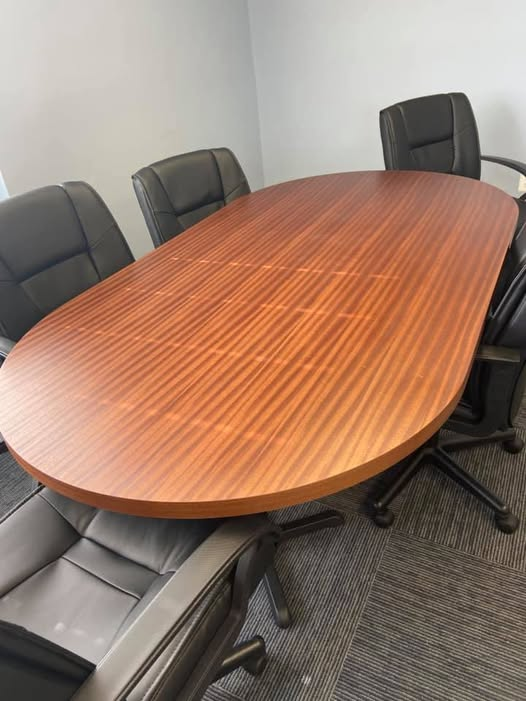 Oval ribbon-striped Sapele mahogany conference table with rich warm tone and matte finish, surrounded by black leather office chairs in a meeting room.