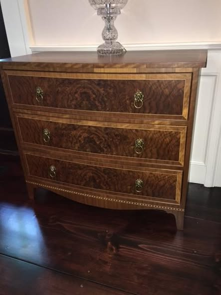 Three-drawer walnut chest with book-matched burl walnut drawer fronts, vertical-grain walnut crossbanding, fine holly stringing, brass ring pulls with lion-head escutcheons, and tapered French feet on a dark plank floor.