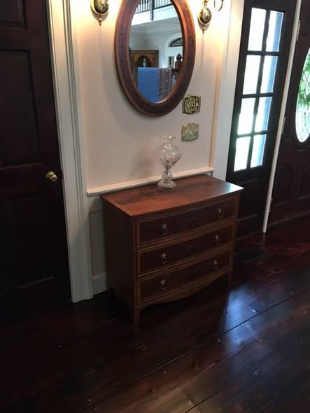 Walnut chest installed in an entry hall beneath an oval burl walnut mirror, with a cut-glass lamp on top and dark wide-plank floors.
