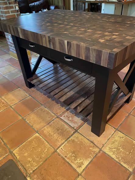 Side view of the walnut butcher-block island showing two drawers with brushed cup pulls, slatted walnut lower shelf, and black X-brace stretchers