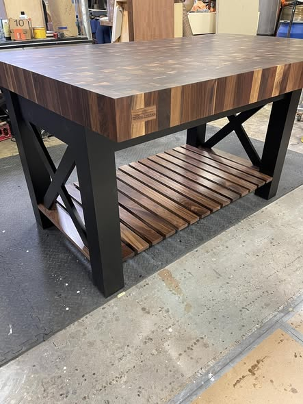 Workshop shot of the finished walnut end-grain island showing the full X-frame base, slatted shelf, and stamped maker's mark on the apron