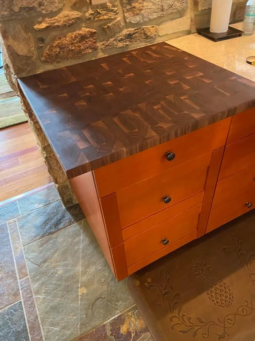 End grain walnut countertop with rich checkerboard grain pattern sitting flush with adjacent stone surface, mounted on an orange-stained cabinet base over slate tile flooring.