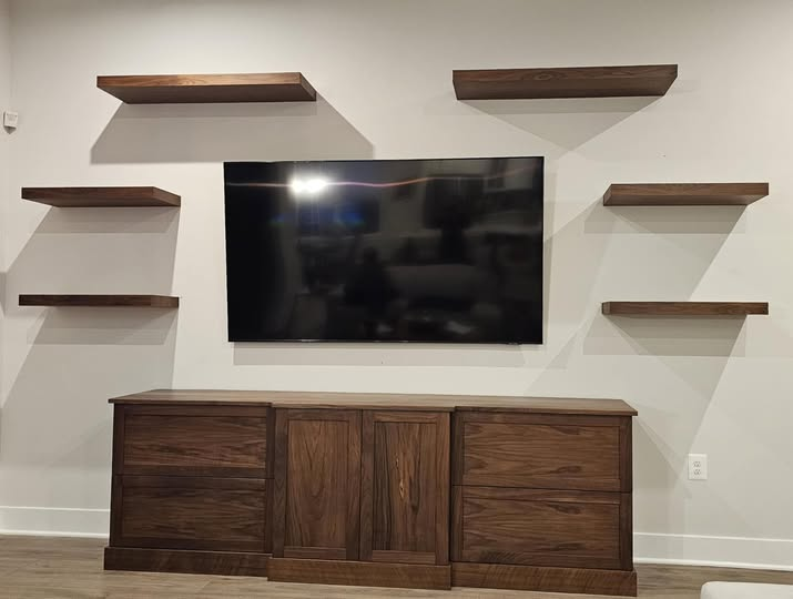 Walnut entertainment center with mounted flat-screen TV, six symmetrical walnut floating shelves flanking the TV, and a long base cabinet with drawers and center doors featuring a bumped-out middle section.