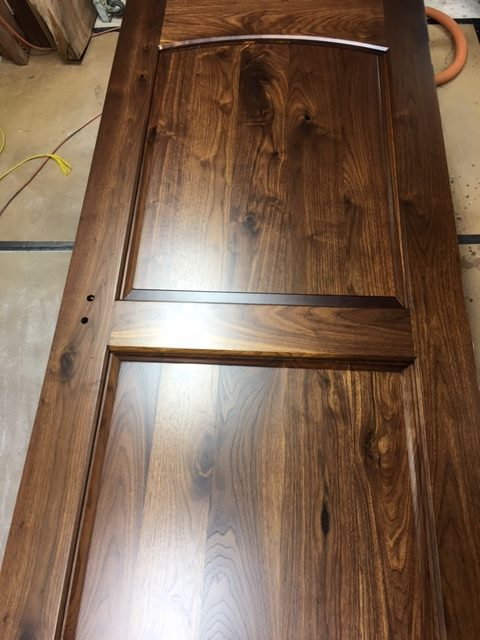 Freshly sprayed solid walnut exterior door laid flat on shop sawhorses, two raised panels with arched top rail, rich chocolate grain and a glossy finish, lockset bores visible in the stile.