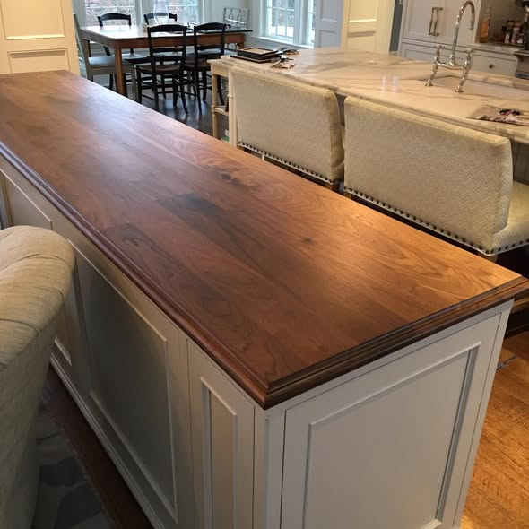 Installed 9-foot long walnut kitchen island top, 28 inches deep, with a shaped edge profile on a soft gray painted island base flanked by upholstered counter stools, marble perimeter counters in the background.