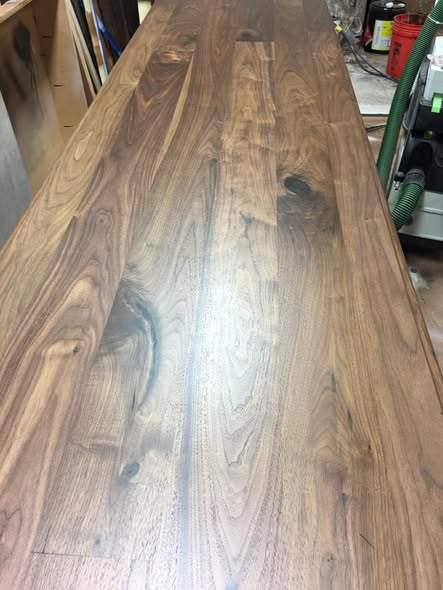 Walnut island top in the shop showing the full length of the wide-board walnut surface with rich grain, mineral streaks, and low-sheen finish.