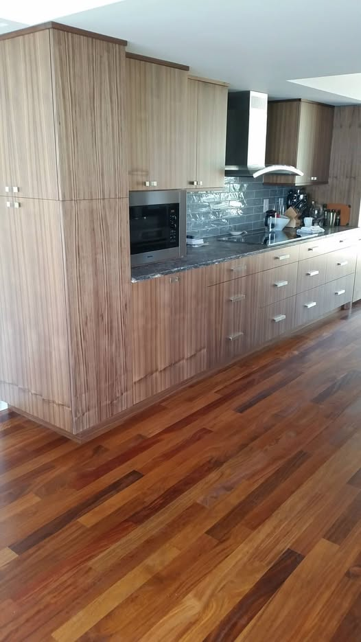 Modern slab-front kitchen in vertical-grain walnut with floor-to-ceiling cabinets, integrated microwave, induction cooktop, stainless wall-mount hood, sculpted silver tile backsplash, and Brazilian cherry hardwood floor